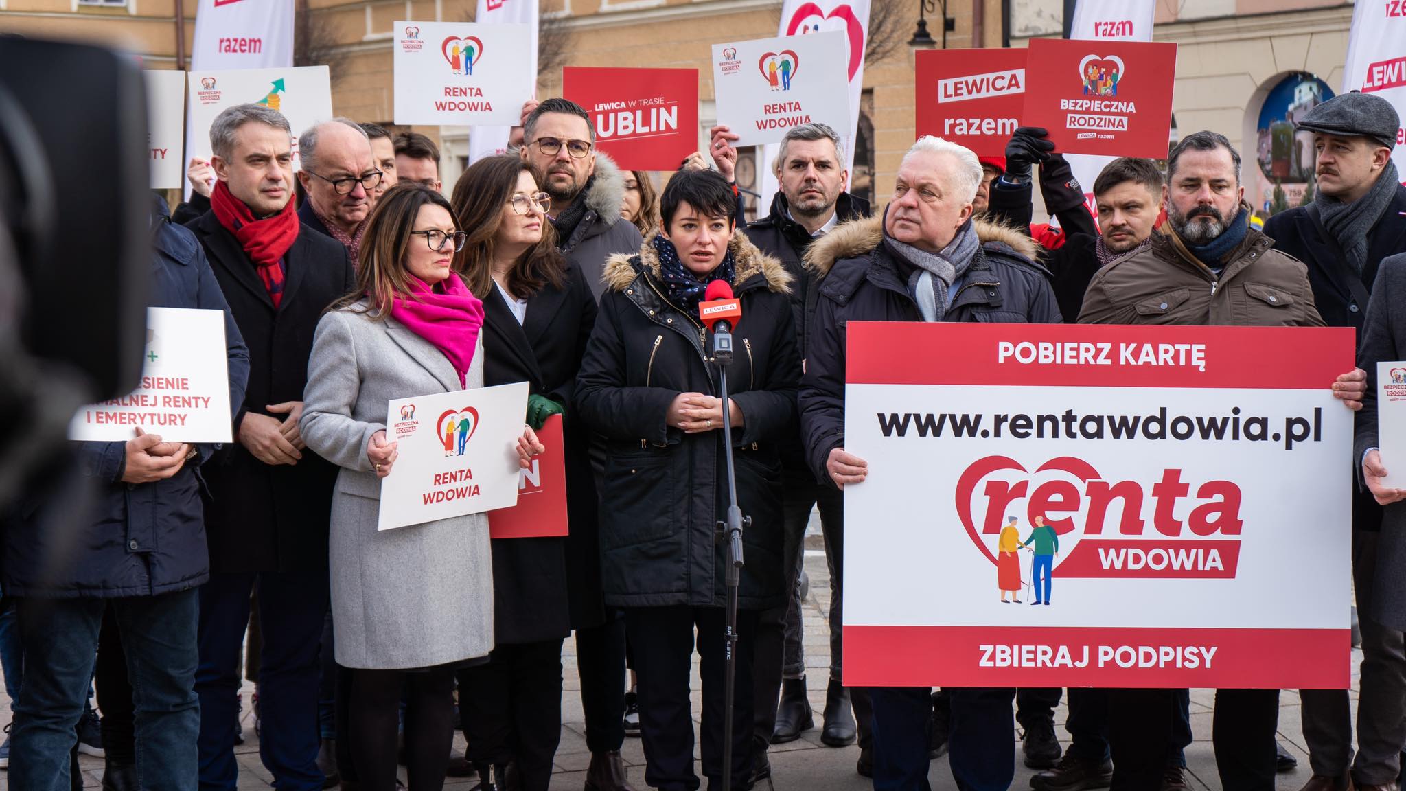 Grupa ludzi na demonstracji na Lubelszczyźnie; kobieta w czarnej kurtce przemawia do mikrofonu, otoczona plakatami „Renta wdowia” i dużym banerem „Pobierz kartę”.