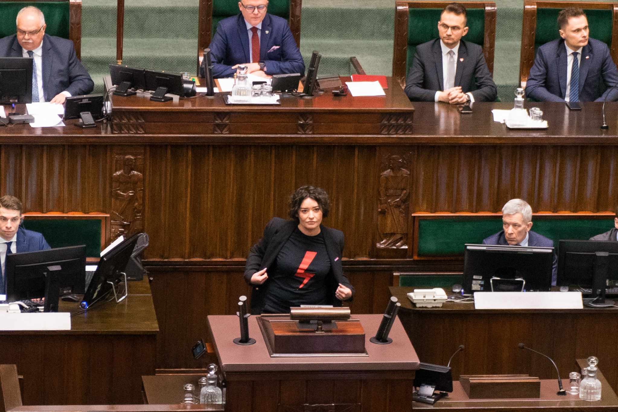 Kobieta w czarnej marynarce stoi przy mównicy w Sejmie, w tle siedzą posłowie przy biurkach z monitorami.