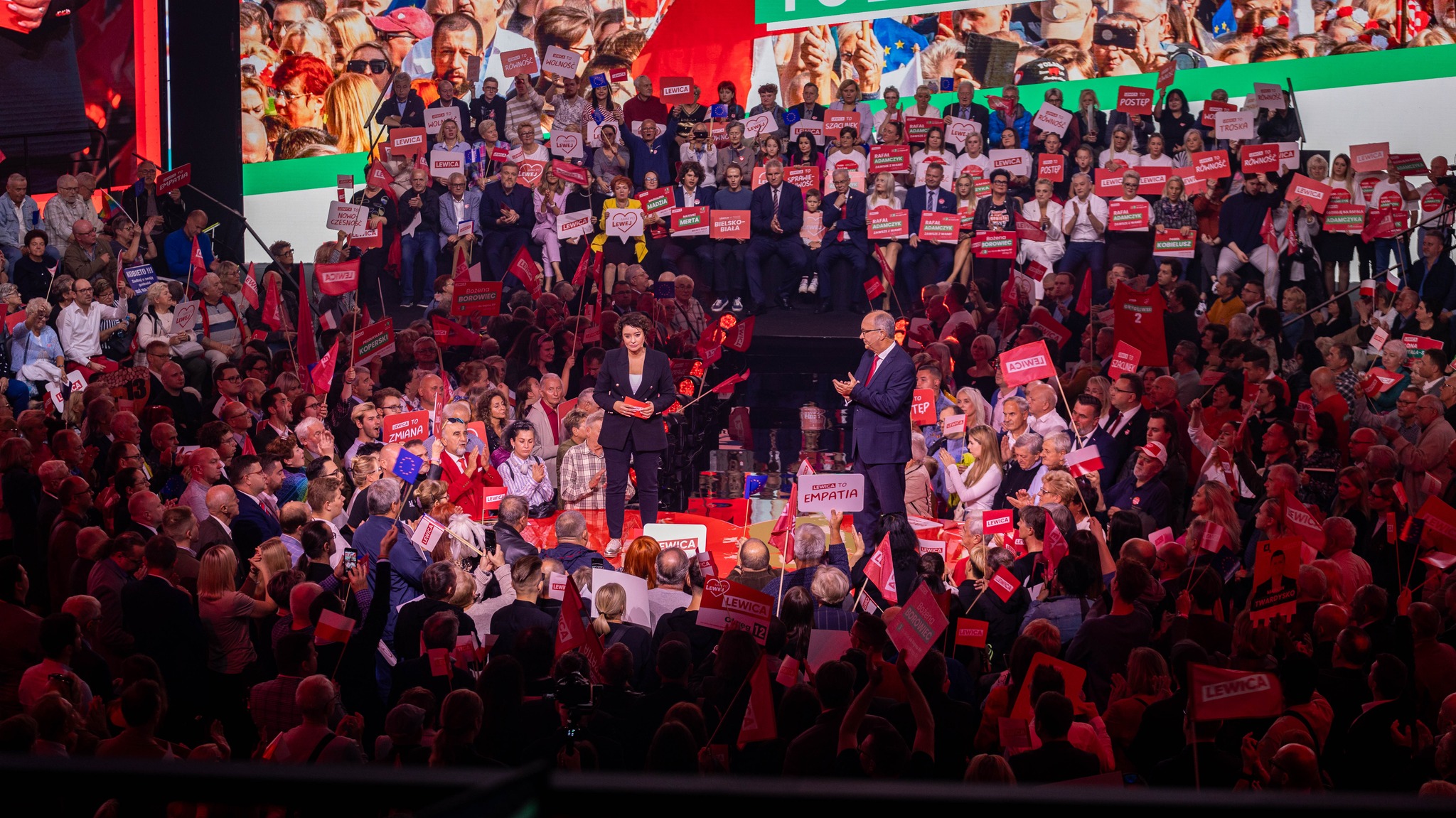 Duża konwencja polityczna z dwoma prowadzącymi na scenie otoczonej tłumem i licznymi czerwonymi plakatami z napisem Lewica.