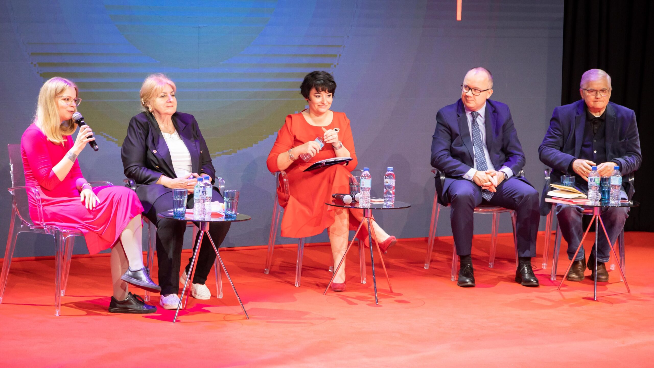 Na zdjęciu przedstawiono pięć osób siedzących na scenie podczas wydarzenia, z których jedna prowadzi rozmowę.