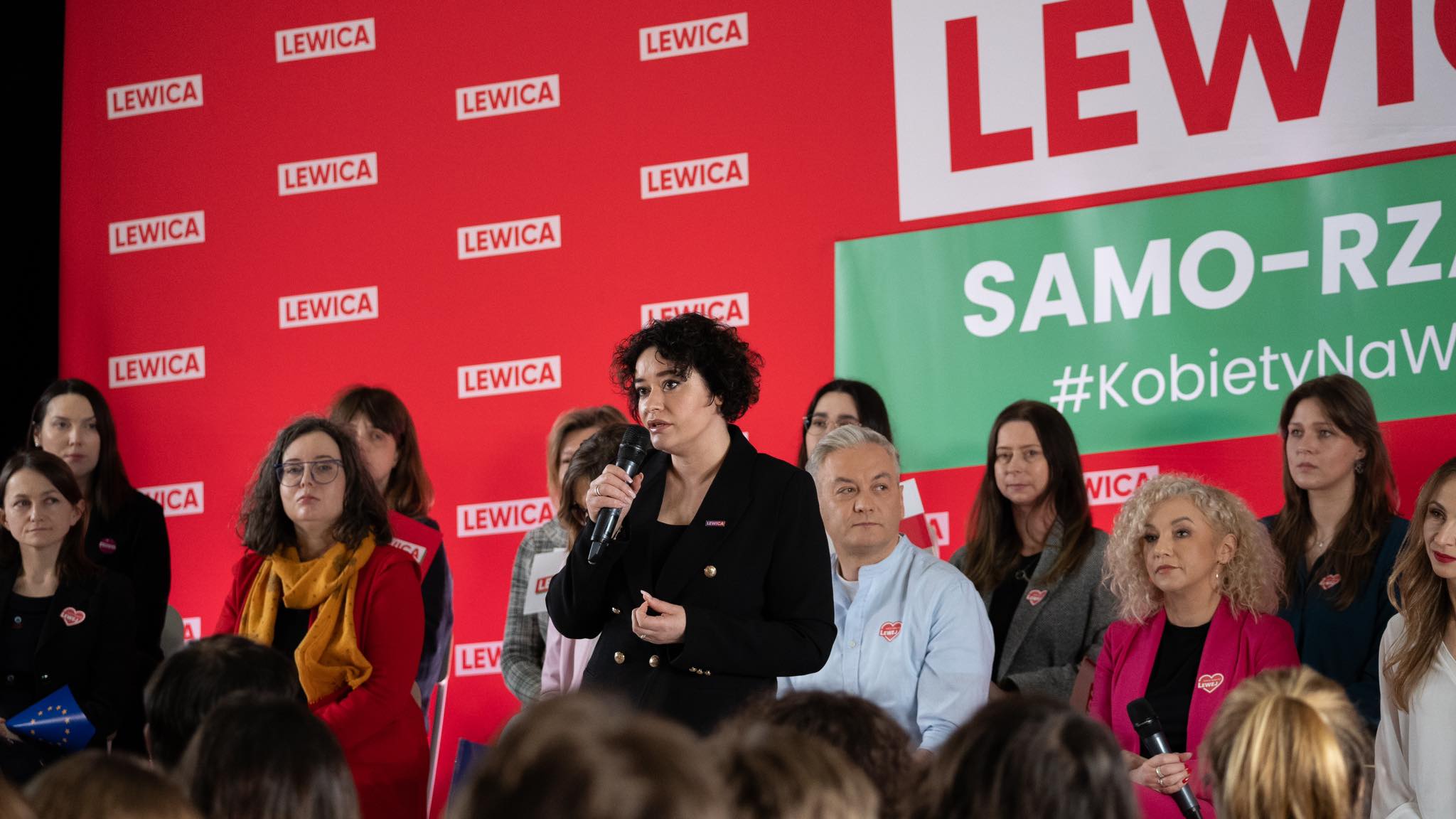 Kandydatki i kandydaci Lewicy przemawiają na scenie podczas konferencji wyborczej, a w tle widoczne są czerwone banery z logiem LEWICA i zielony baner.