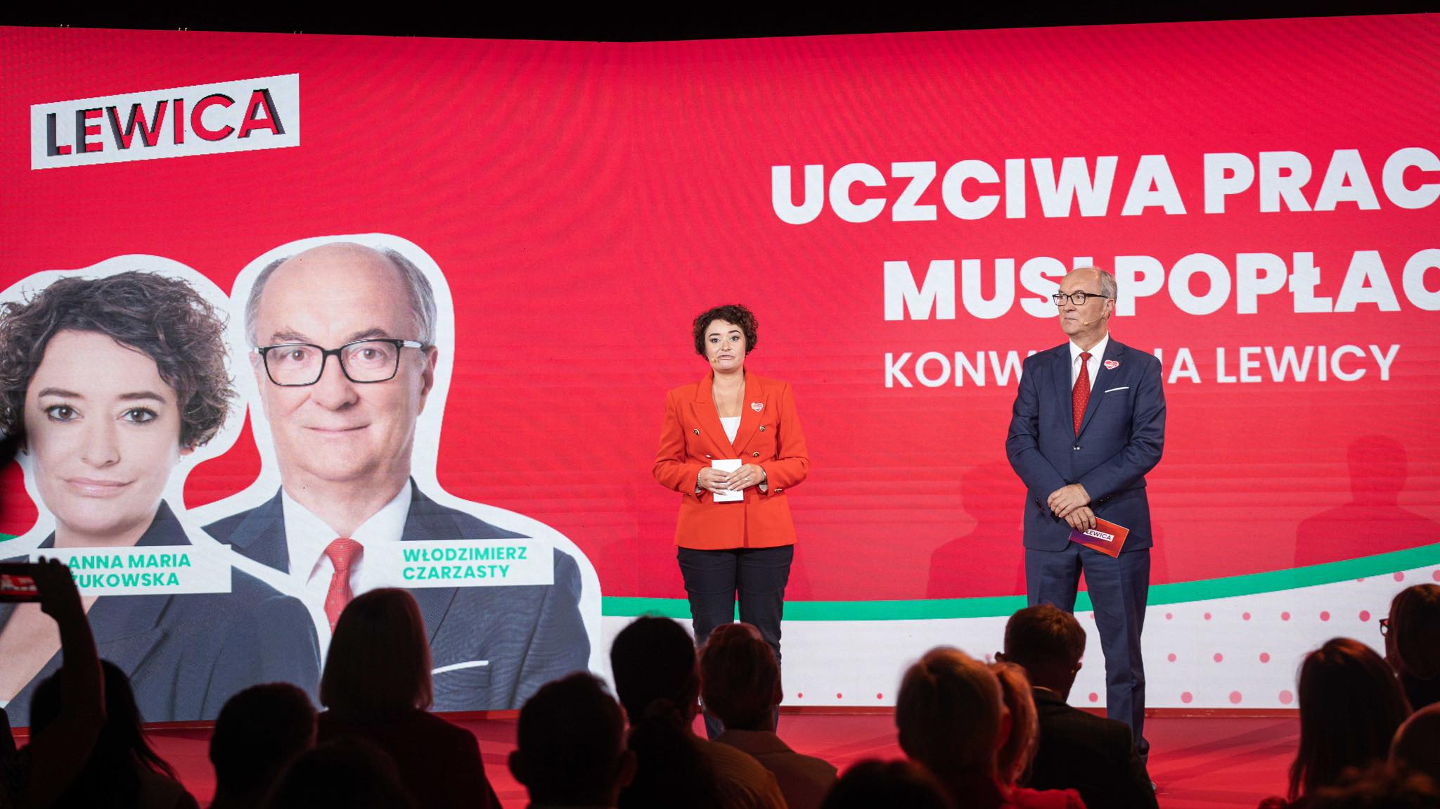 Scena polityczna na czerwonym tle z hasłem Lewica „Uczciwa praca musi popłacać”, dwie osoby stojące na scenie obok dużych plakatów z wizerunkami kandydatów.