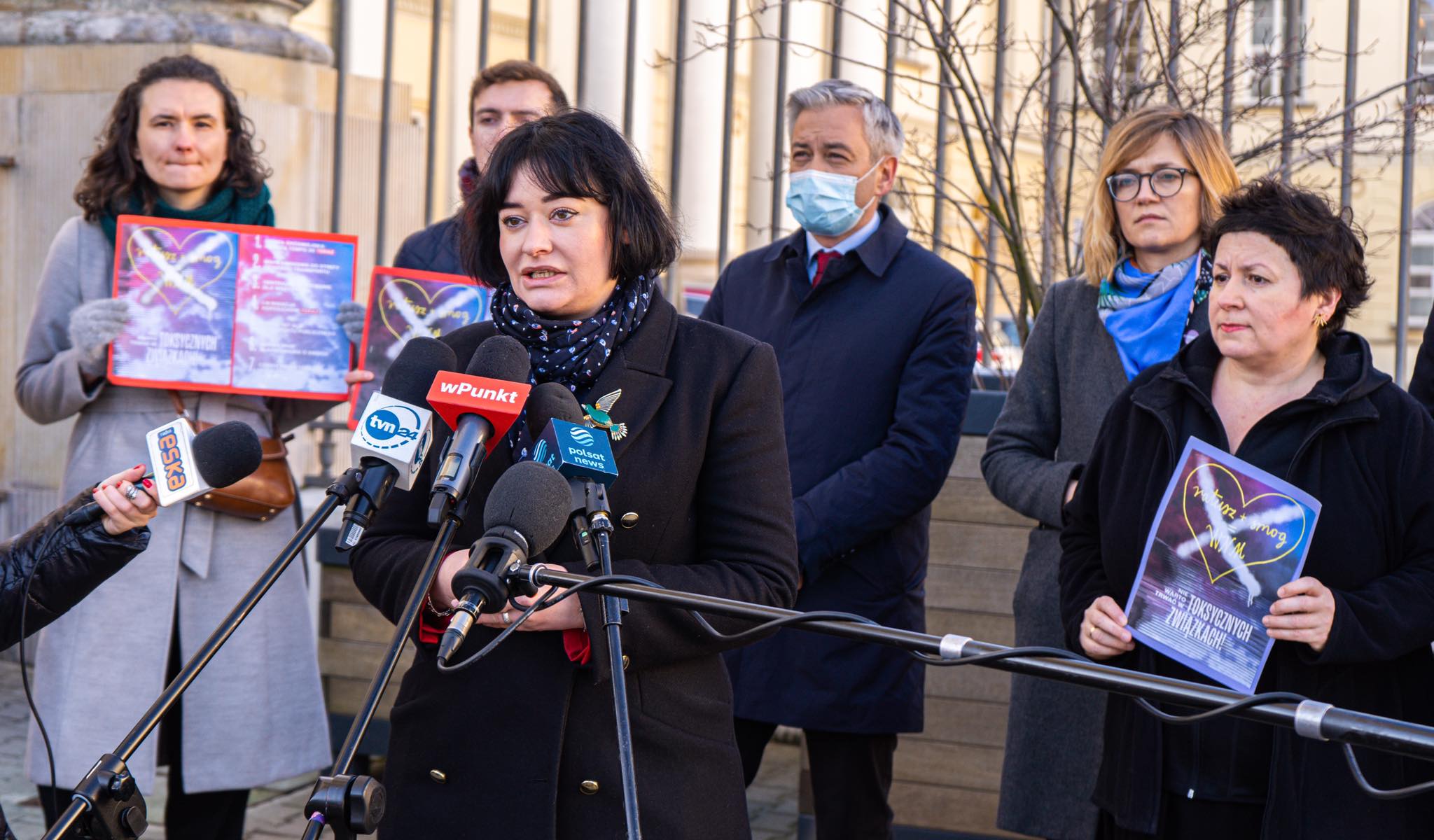Na zdjęciu grupa osób uczestniczy w konferencji prasowej, podczas której mówcy trzymają plakaty i mówią do mikrofonów.