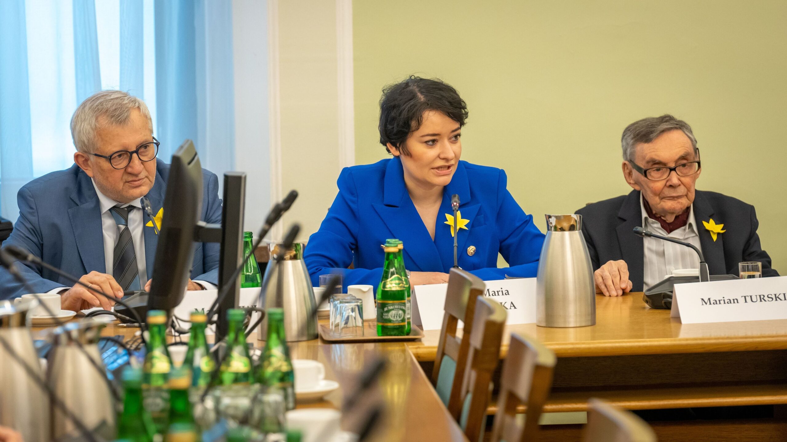 Na zdjęciu widać trzech uczestników spotkania przy stole, na którym stoją butelki wody i filiżanki; w centrum siedzi kobieta w niebieskiej marynarce, a obok niej dwóch mężczyzn.