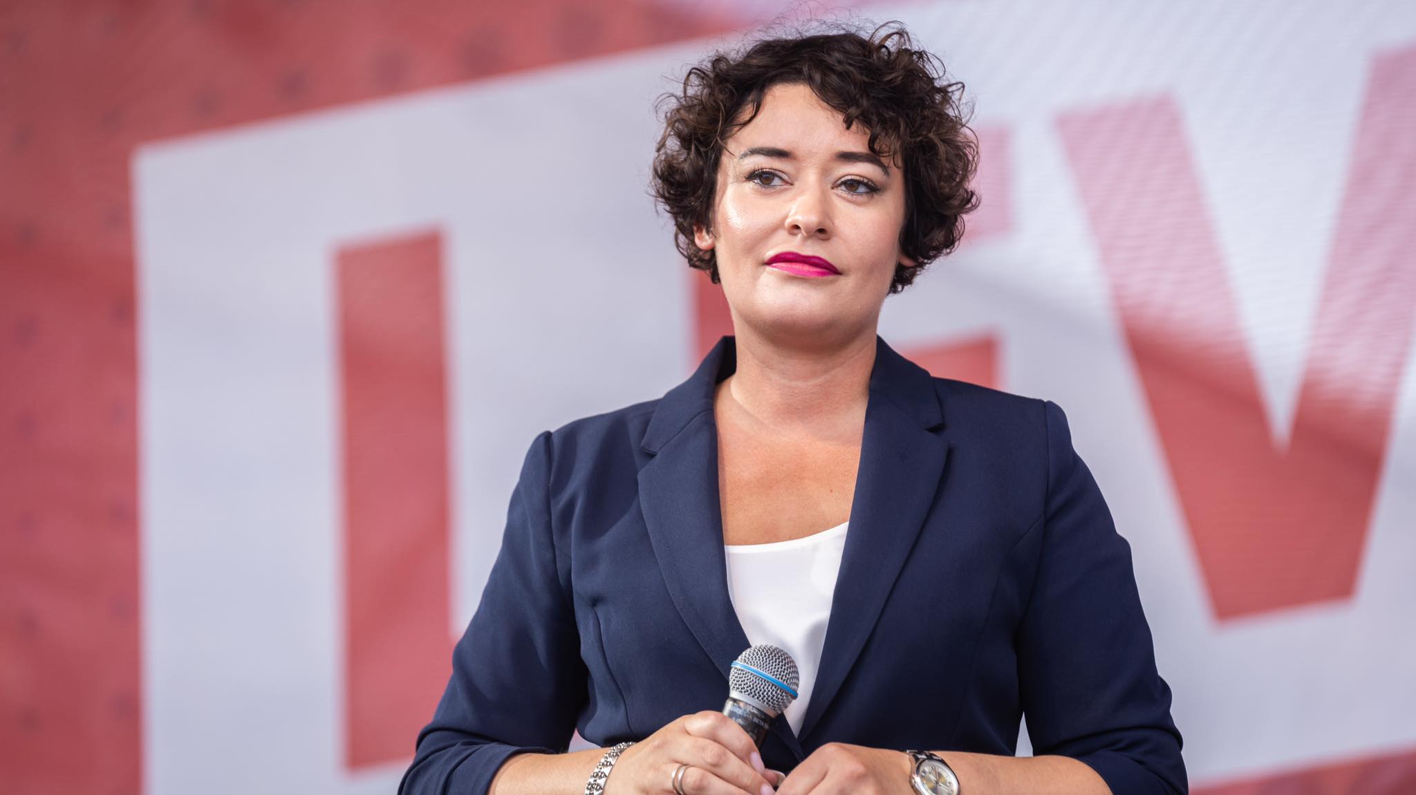 Kobieta w granatowej marynarce trzyma mikrofon podczas wiecu wyborczego w Częstochowie, na tle czerwono-białego baneru.