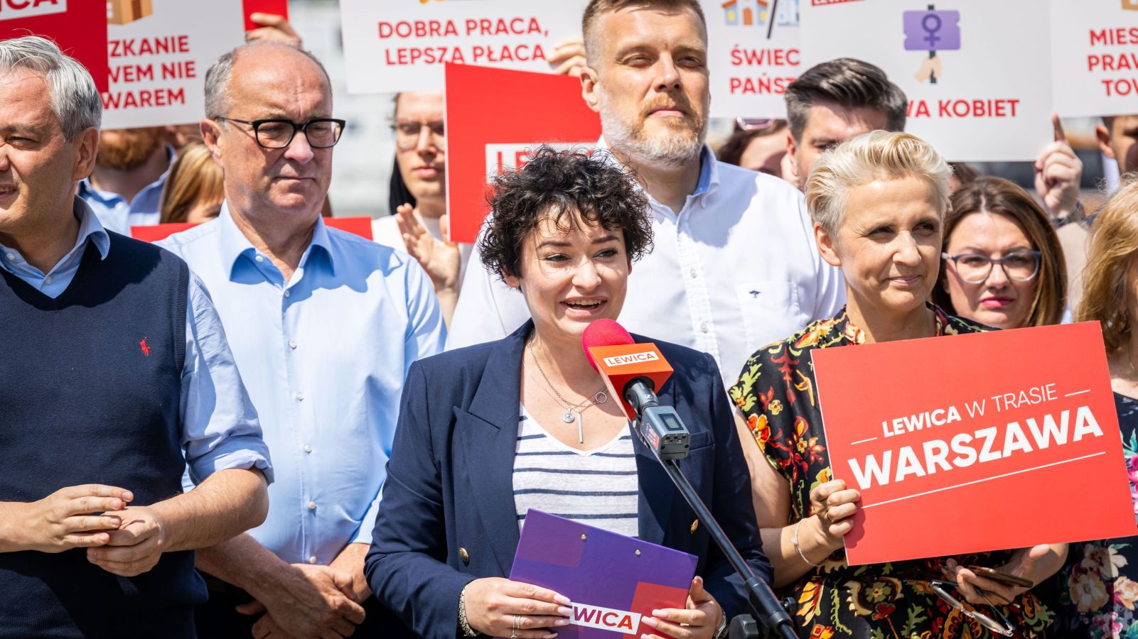 Kobieta przemawia do mikrofonu podczas konferencji w ramach Wakacyjnej Trasy Lewicy, otoczona członkami partii trzymającymi czerwone plakaty z hasłami.