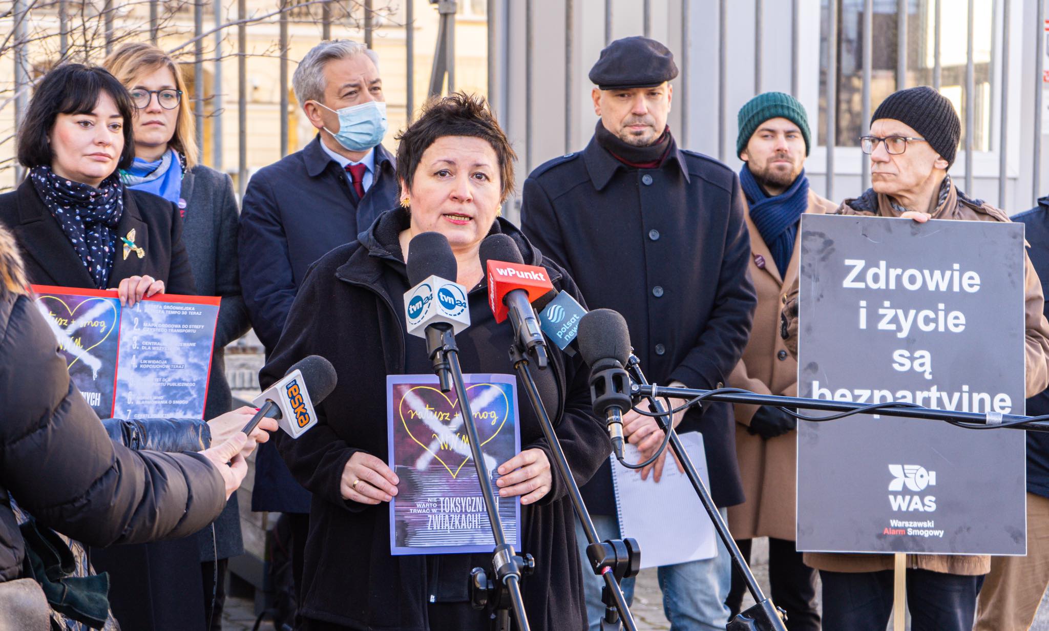 Kobieta wygłasza przemówienie do mediów podczas konferencji prasowej w Warszawie na temat czystego powietrza, otoczona dziennikarzami i innymi uczestnikami z plakatami.