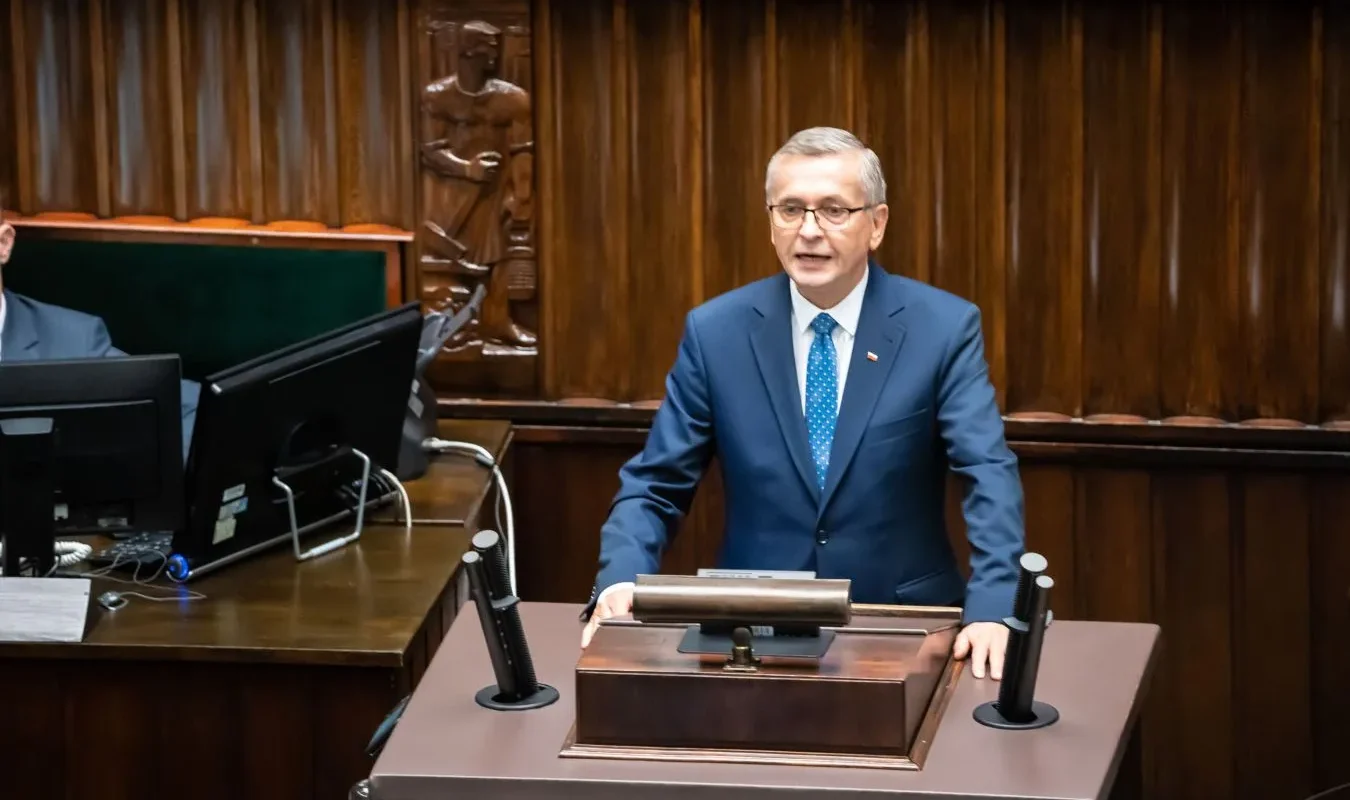 Mężczyzna w granatowym garniturze stoi przy mównicy w sali plenarnej i wygłasza przemówienie.