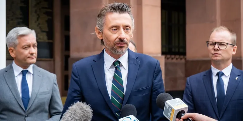 Trzej mężczyźni w garniturach stoją przed budynkiem, jeden z nich przemawia do mediów, a pozostali go słuchają.