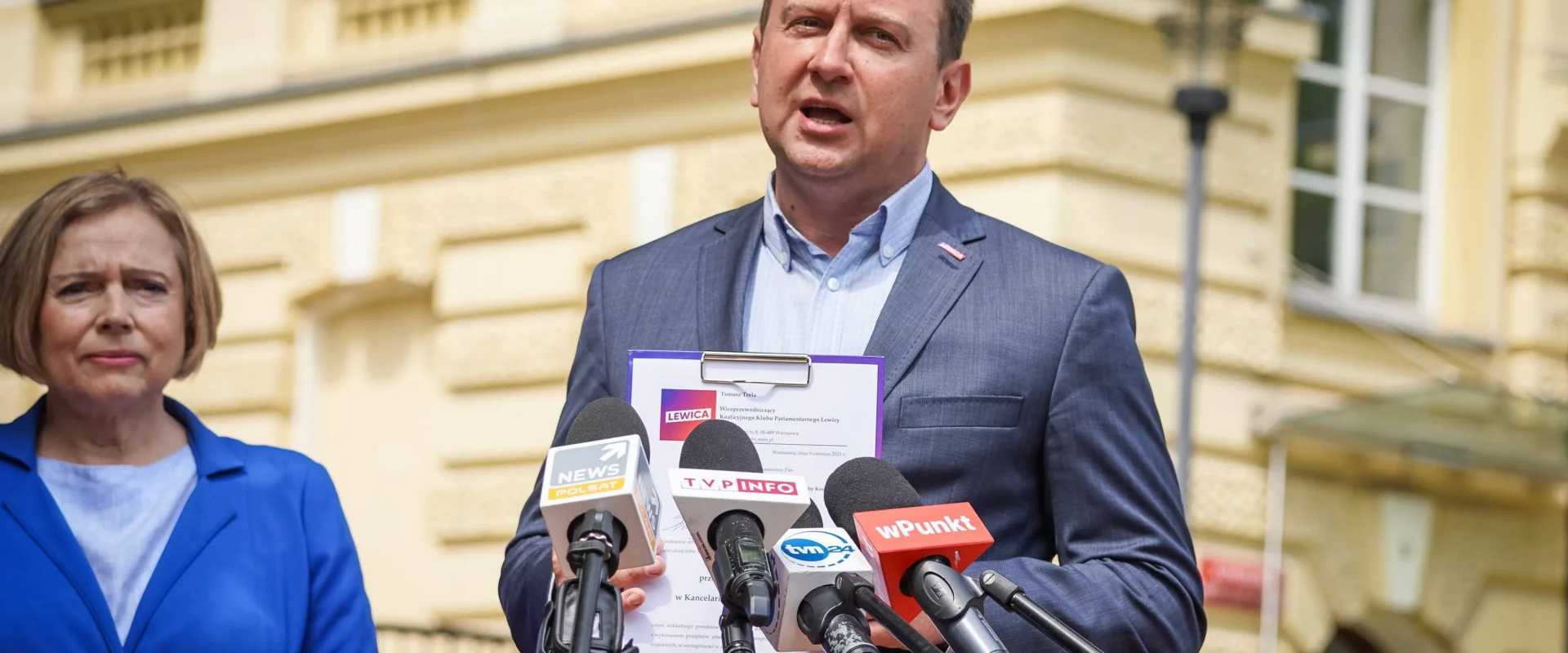 Polityk w granatowym garniturze przemawia do mediów na zewnątrz budynku, trzymając plik z dokumentem dotyczącym wniosku do NIK o kontrolę obiegu dokumentów elektronicznych w KPRM, a obok stoi kobieta w niebieskim żakiecie.