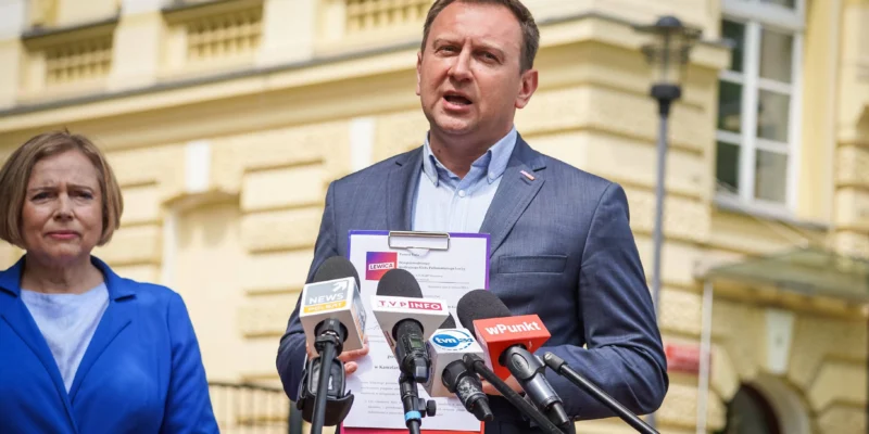 Polityk w granatowym garniturze przemawia do mediów na zewnątrz budynku, trzymając plik z dokumentem dotyczącym wniosku do NIK o kontrolę obiegu dokumentów elektronicznych w KPRM, a obok stoi kobieta w niebieskim żakiecie.