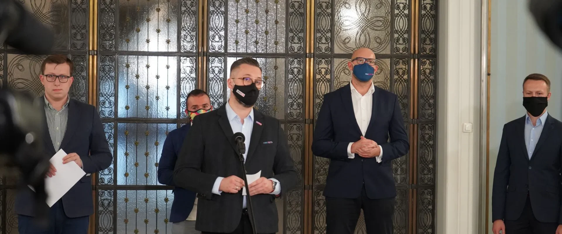 Mężczyzna w ciemnym garniturze i czarnej maseczce przemawia do mikrofonu podczas konferencji prasowej, otoczony innymi mężczyznami w garniturach, przed ozdobnymi, metalowymi drzwiami.