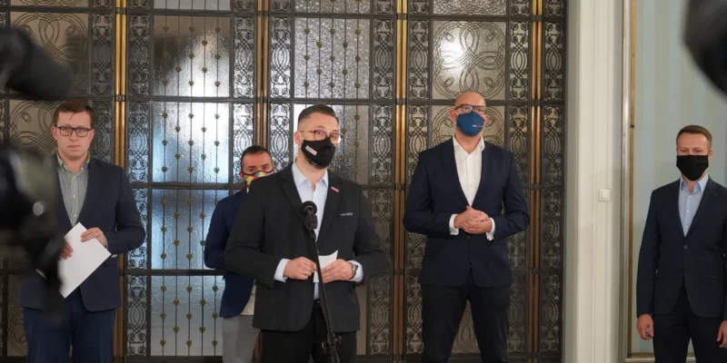 Mężczyzna w ciemnym garniturze i czarnej maseczce przemawia do mikrofonu podczas konferencji prasowej, otoczony innymi mężczyznami w garniturach, przed ozdobnymi, metalowymi drzwiami.