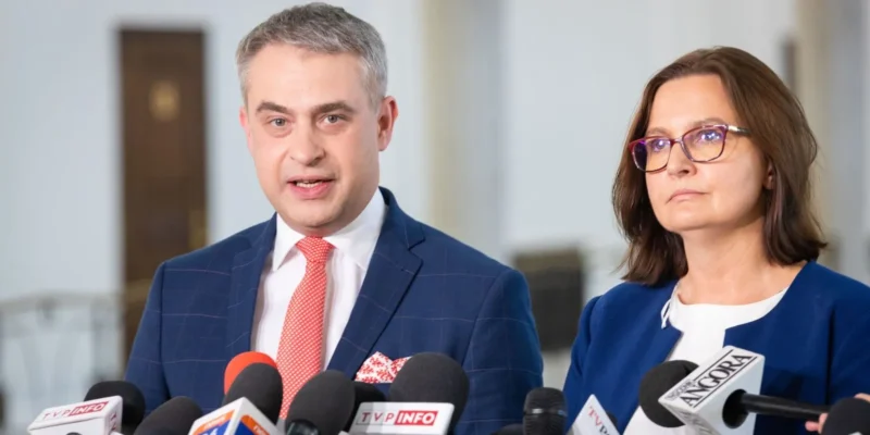 Dwóch polityków stoi przy licznym zestawie mikrofonów podczas konferencji prasowej w budynku; mężczyzna w granatowym garniturze z czerwonym krawatem przemawia, obok niego stoi kobieta w niebieskim żakiecie.