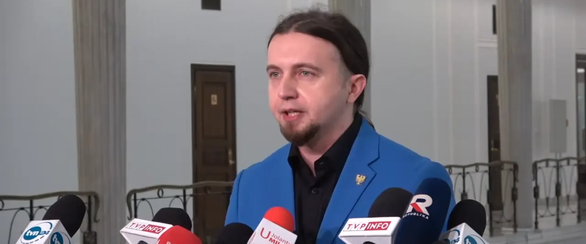 Mężczyzna w niebieskim garniturze przemawia do dziennikarzy, otoczony mikrofonami w budynku użyteczności publicznej.