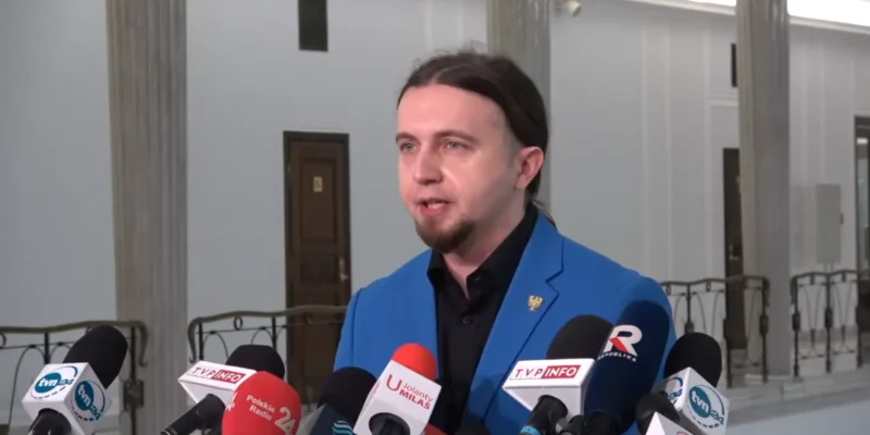 Mężczyzna w niebieskim garniturze przemawia do dziennikarzy, otoczony mikrofonami w budynku użyteczności publicznej.