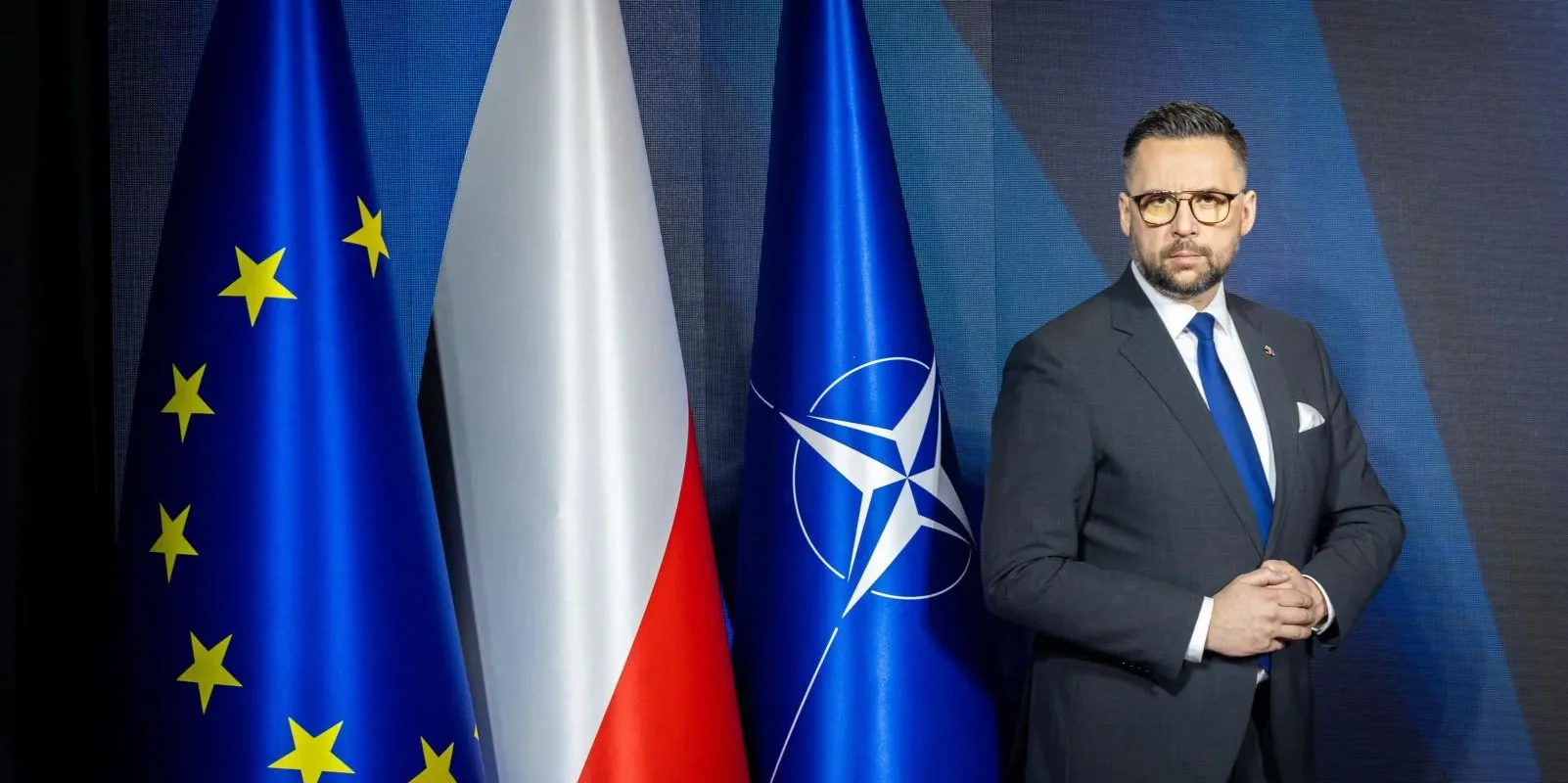 Mężczyzna w garniturze stoi na tle flag Unii Europejskiej, Polski i NATO.