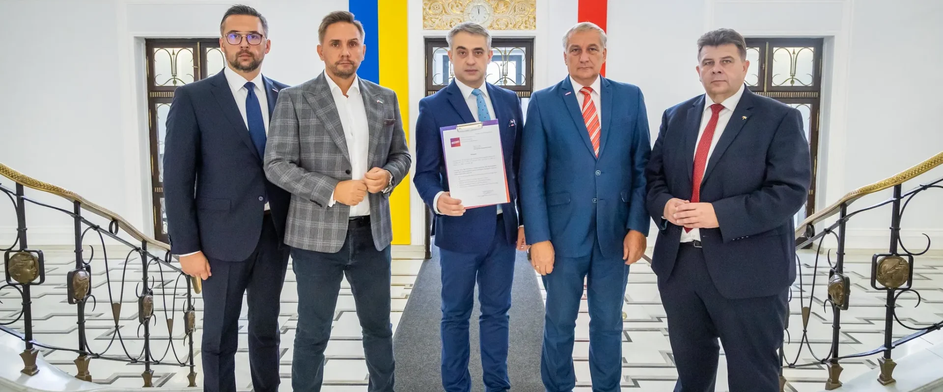 Grupa pięciu mężczyzn w garniturach stoi w eleganckim wnętrzu, trzymając dokumenty, z flagami w tle.