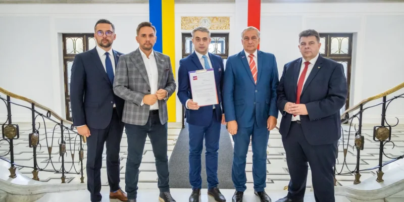 Grupa pięciu mężczyzn w garniturach stoi w eleganckim wnętrzu, trzymając dokumenty, z flagami w tle.