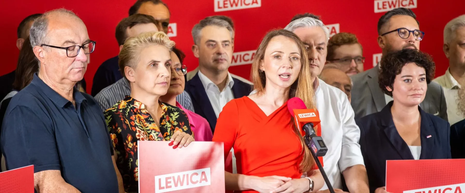 Kobieta w czerwonej sukience wygłasza przemówienie do mikrofonu podczas konferencji partii Lewica, otoczona innymi politykami na czerwonym tle z logami Lewica.