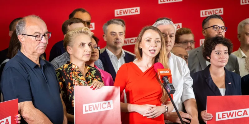 Kobieta w czerwonej sukience wygłasza przemówienie do mikrofonu podczas konferencji partii Lewica, otoczona innymi politykami na czerwonym tle z logami Lewica.