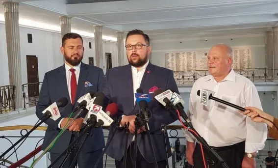 Trzech mężczyzn stoi przy pulpicie z wieloma mikrofonami podczas konferencji prasowej w reprezentacyjnym holu, przemawiając o podniesieniu cen paliw i wzroście kosztów życia.