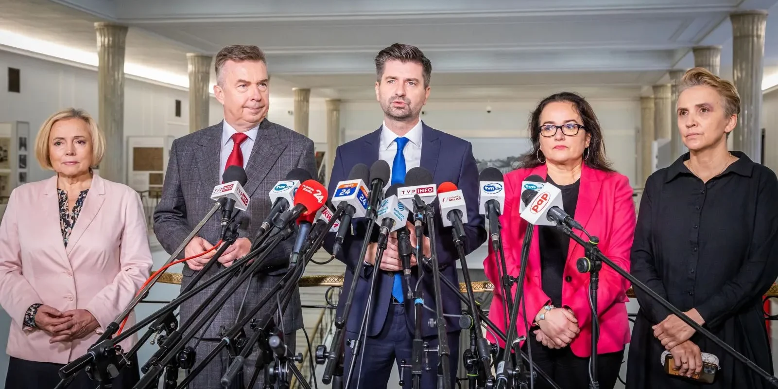 Na zdjęciu widoczni są pięciu polityków stojących przed mikrofonami podczas konferencji prasowej.