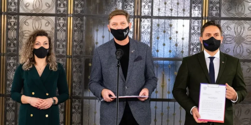 Trzy osoby w maseczkach stoją przed ozdobnymi drzwiami z metalowymi wzorami; pośrodku mężczyzna przy mikrofonie trzyma dokument, po lewej kobieta w zielonej sukience, a po prawej mężczyzna w garniturze trzymający kolejny dokument.