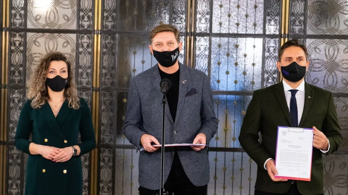 Trzy osoby w maseczkach stoją przed ozdobnymi drzwiami z metalowymi wzorami; pośrodku mężczyzna przy mikrofonie trzyma dokument, po lewej kobieta w zielonej sukience, a po prawej mężczyzna w garniturze trzymający kolejny dokument.