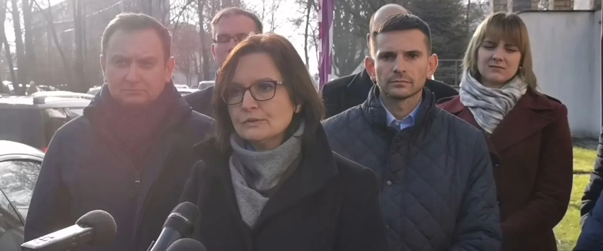 Grupa osób stojąca na zewnątrz podczas konferencji prasowej; kobieta w okularach przemawia do mikrofonów.