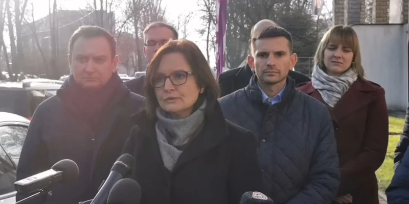 Grupa osób stojąca na zewnątrz podczas konferencji prasowej; kobieta w okularach przemawia do mikrofonów.