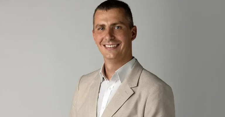 Portret mężczyzny w beżowym garniturze na neutralnym tle, uśmiechnięty do kamery.