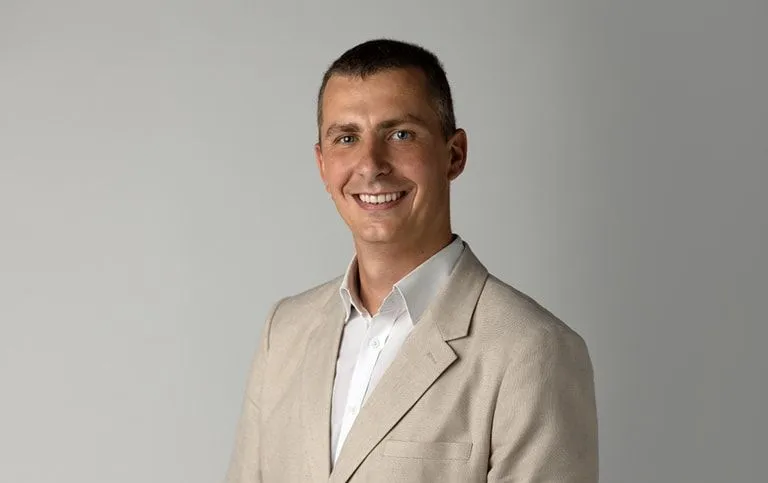 Portret mężczyzny w beżowym garniturze na neutralnym tle, uśmiechnięty do kamery.