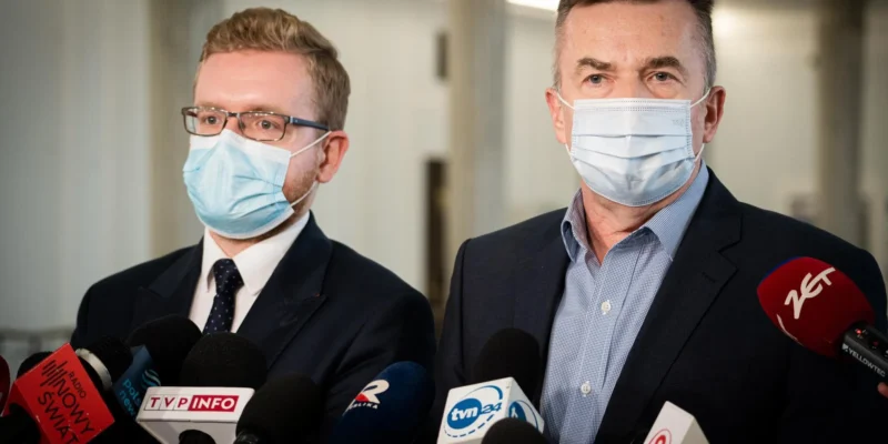 Na zdjęciu widać dwóch mężczyzn w maseczkach, stojących przed mikrofonami i udzielających wywiadu.