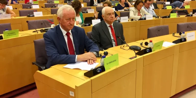 Uczestnicy debaty na temat stanu praworządności w Polsce w Parlamencie Europejskim siedzą w sali obrad; na pierwszym planie dwóch mężczyzn w garniturach przy stolikach z mikrofonami i dokumentami.