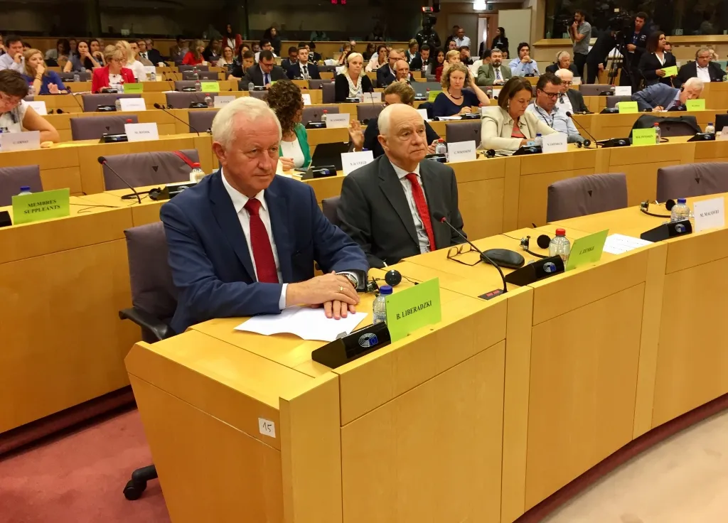 Uczestnicy debaty na temat stanu praworządności w Polsce w Parlamencie Europejskim siedzą w sali obrad; na pierwszym planie dwóch mężczyzn w garniturach przy stolikach z mikrofonami i dokumentami.