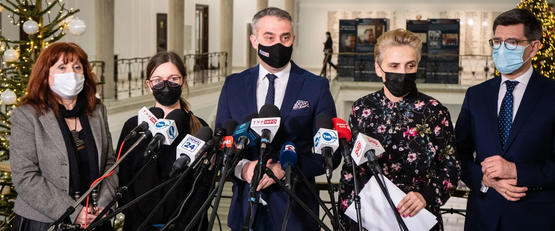 Pięć osób w maseczkach stoi na konferencji prasowej w holu z choinką w tle, otoczone licznymi mikrofonami z logotypami mediów.