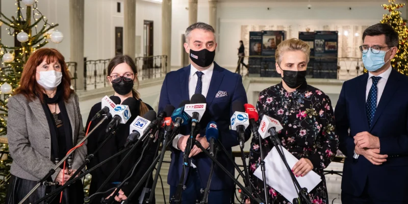 Pięć osób w maseczkach stoi na konferencji prasowej w holu z choinką w tle, otoczone licznymi mikrofonami z logotypami mediów.
