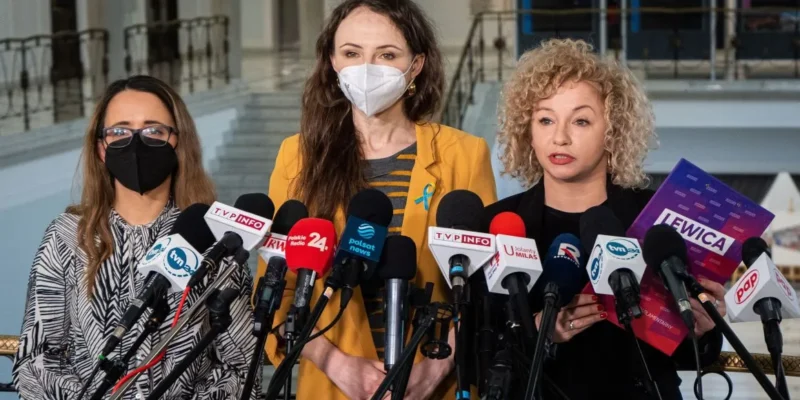 Trzy kobiety stoją przed mikrofonami na konferencji prasowej, prezentując materiały związane z ich działalnością.