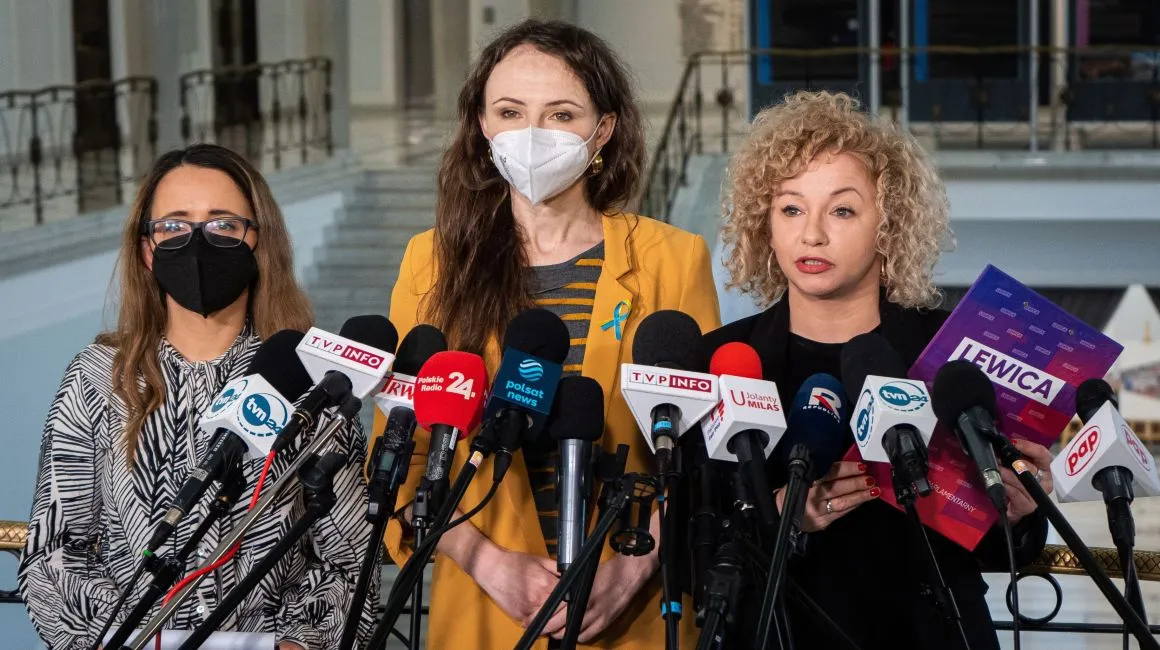 Trzy kobiety stoją przed mikrofonami na konferencji prasowej, prezentując materiały związane z ich działalnością.