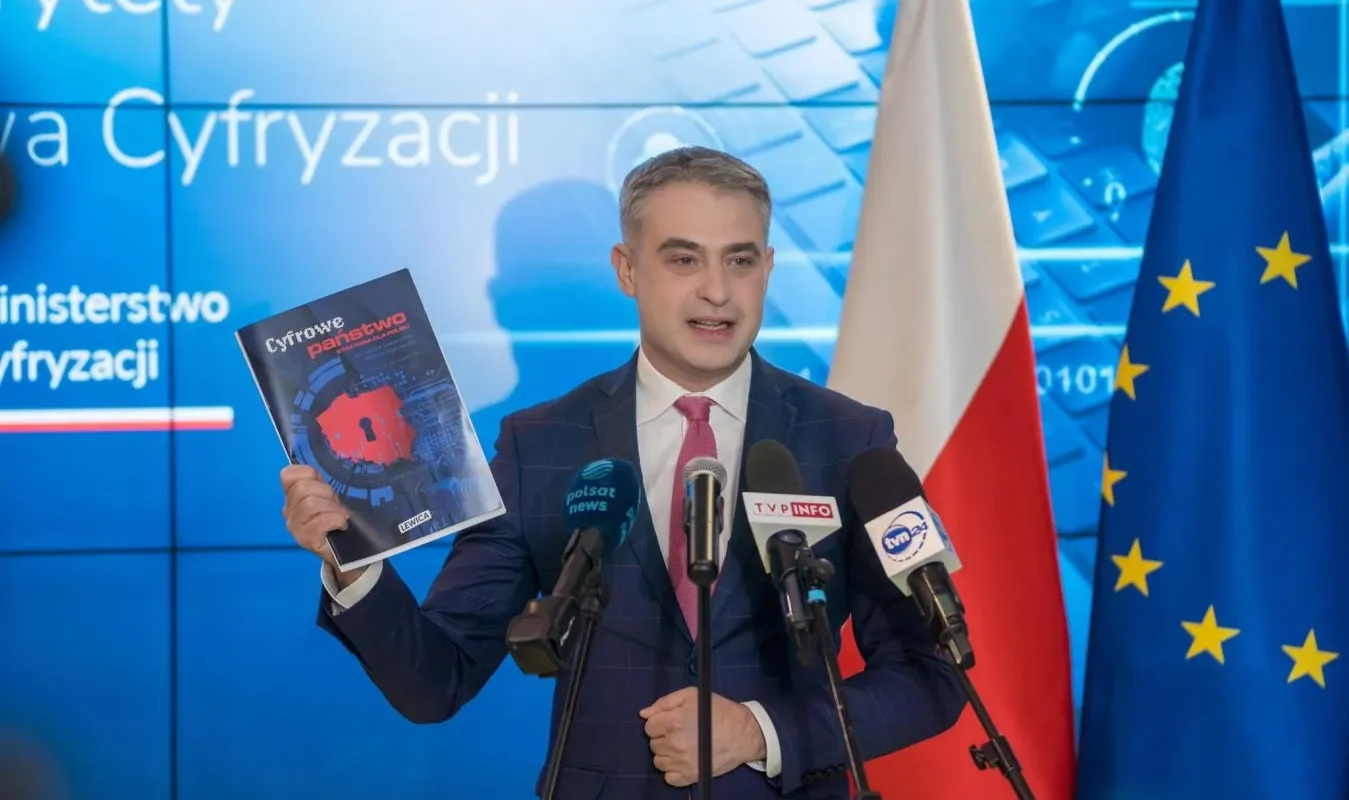 Mężczyzna w garniturze podczas konferencji prasowej trzyma w lewej ręce materiał zatytułowany „Cyfrowe państwo”, stoi przy mikrofonach, z flagą Polski i flagą Unii Europejskiej w tle.