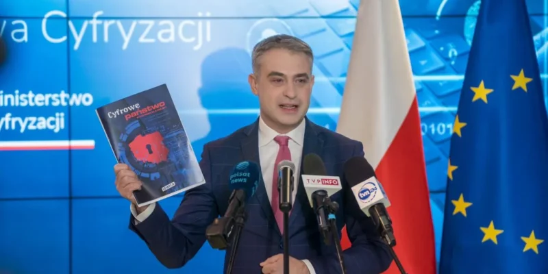 Mężczyzna w garniturze podczas konferencji prasowej trzyma w lewej ręce materiał zatytułowany „Cyfrowe państwo”, stoi przy mikrofonach, z flagą Polski i flagą Unii Europejskiej w tle.