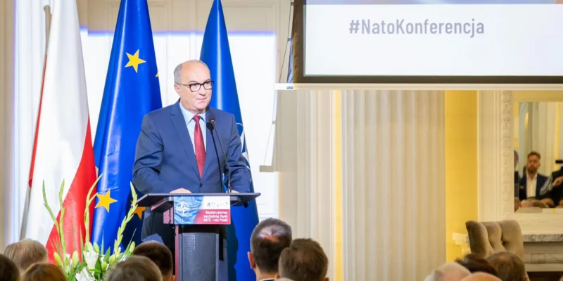 Na zdjęciu widoczny jest mężczyzna stojący przy mównicy podczas konferencji dotyczącej NATO, z flagami Polski, UE oraz NATO w tle.