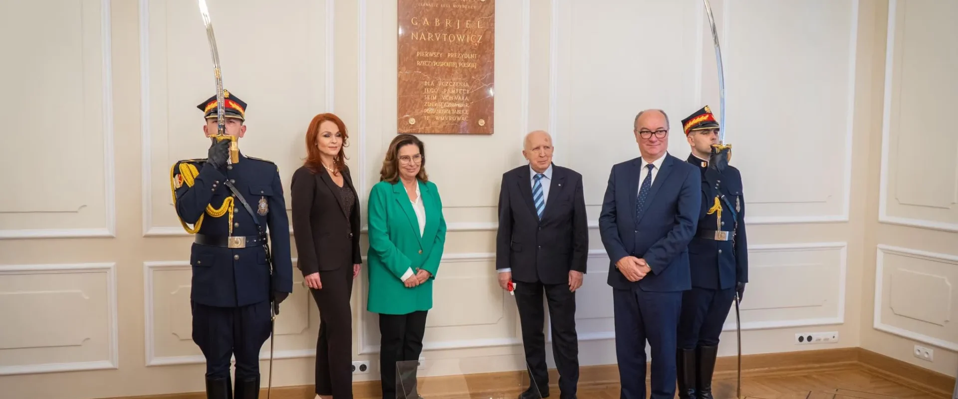 Na zdjęciu znajduje się grupa osób stojących przed tablicą upamiętniającą Gabriela Narutowicza, z żołnierzami w mundurach stojącymi po bokach.