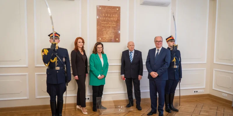 Na zdjęciu znajduje się grupa osób stojących przed tablicą upamiętniającą Gabriela Narutowicza, z żołnierzami w mundurach stojącymi po bokach.