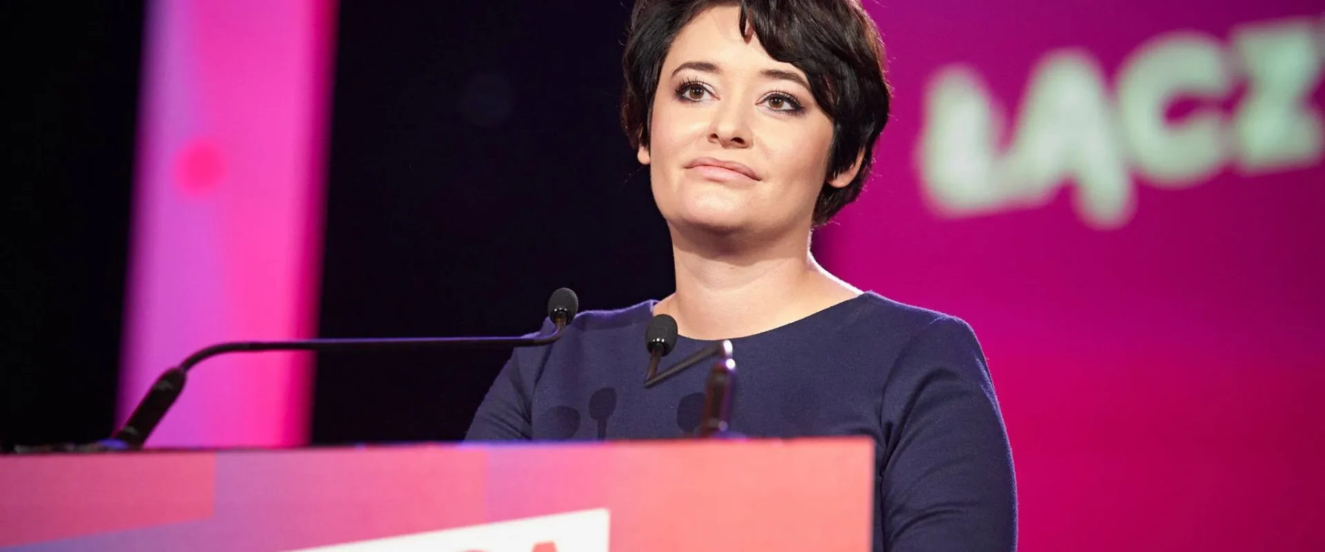 Kobieta z krótkimi włosami w granatowej sukience stoi przy mównicy z logo partii Lewica, podczas wystąpienia na scenie z kolorowym tłem.