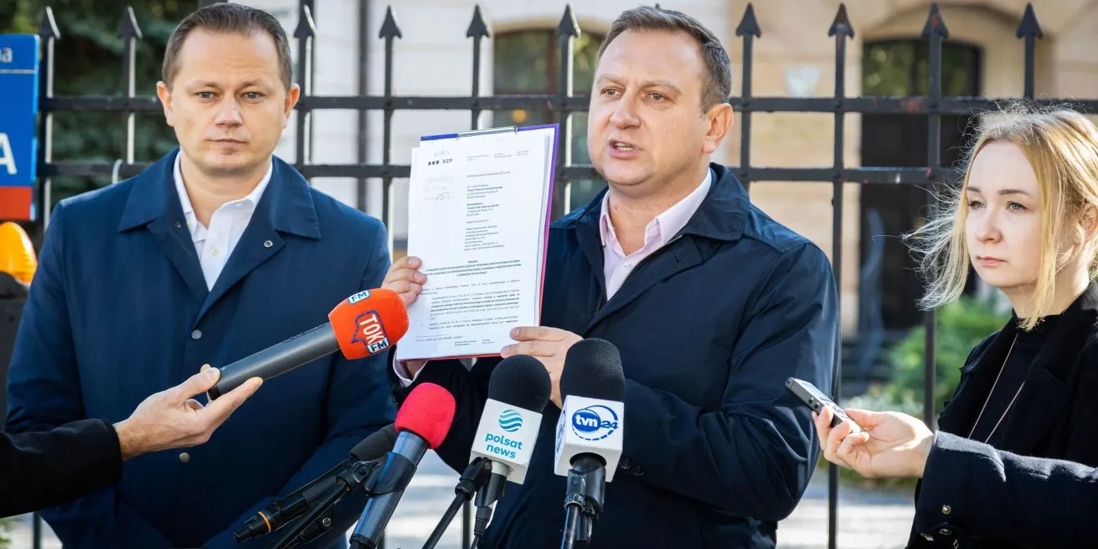 Na zdjęciu widać mężczyznę trzymającego dokument, otoczonego przez dziennikarzy i znaczniki mikrofonów, podczas konferencji prasowej.