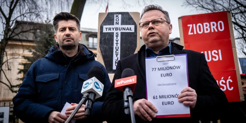 Dwóch mężczyzn podczas konferencji prasowej stoi przy mikrofonach, w tle widoczne są plakaty i banery związane z Europejską Konwencją Praw Człowieka, w tym z napisem Ziobro oraz kartką z informacją o 77 milionach euro.