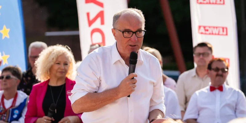 Mężczyzna w białej koszuli i czarnych okularach przemawia do zgromadzonych, trzymając mikrofon, na tle bannerów i publiczności.