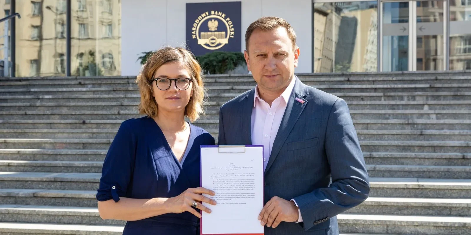 Na zdjęciu widoczni są dwaj ludzie pozujący na schodach przed budynkiem Narodowego Banku Polskiego, trzymający dokument.